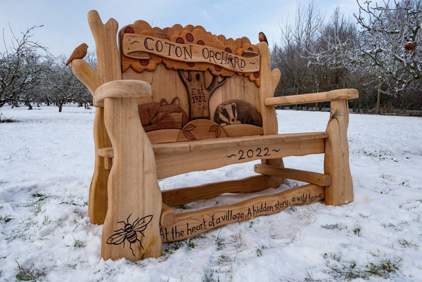 Handcrafted wooden bench with intricate carvings of a badger, tree, and bee, featuring the inscription 'Coton Orchard - Est. 1922' and '2022.' Set in a snowy orchard