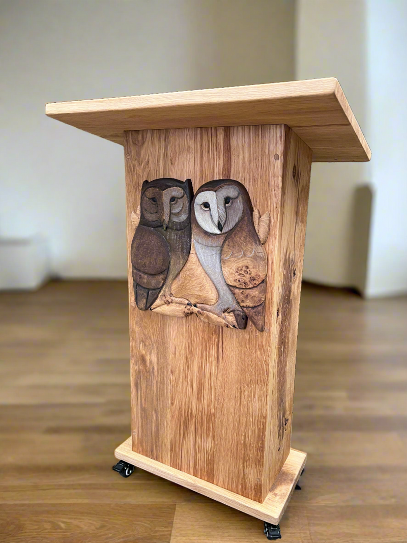 Front view of a handcrafted oak reading lectern with wheels, featuring two intricately carved owls on the front. The lectern is positioned in a workshop environment with various woodworking tools and materials visible in the background