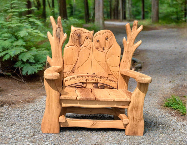 Handcrafted wooden memorial bench with two detailed owl carvings on the backrest. The bench is engraved with 'In loving memory of Graham Peter Bartram 1949-2023' and is set along a forest path, surrounded by greenery