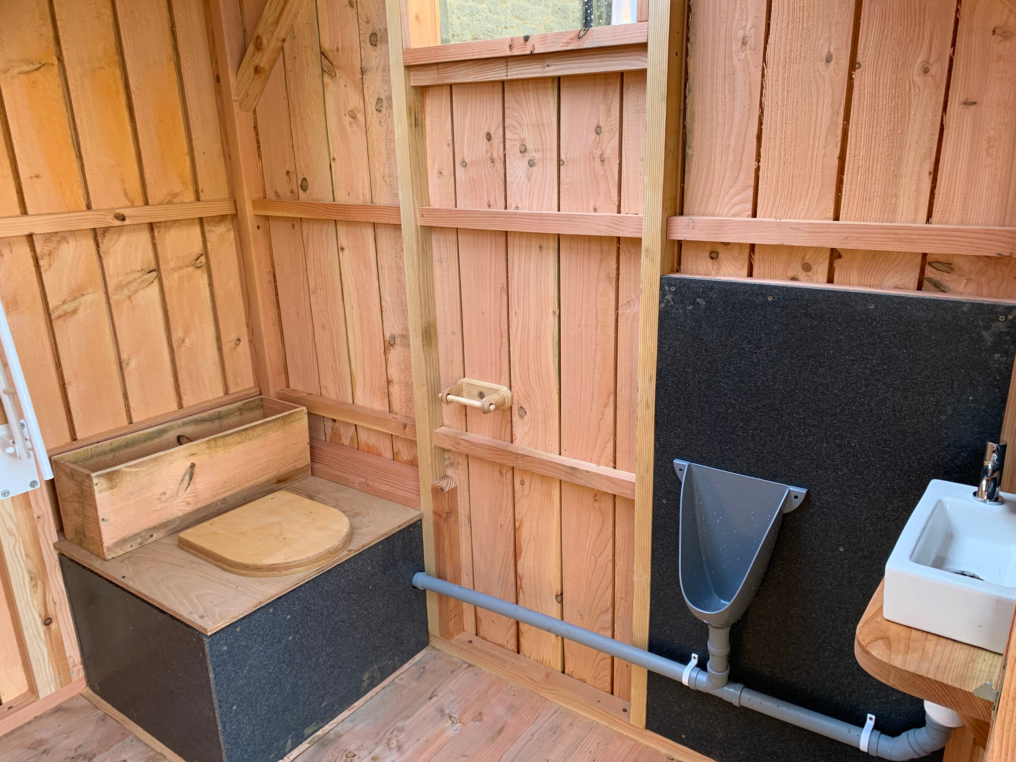 Interior view of a compost toilet with wooden seat, urinal, and handwashing sink, designed by Free Range Designs for sustainable sanitation