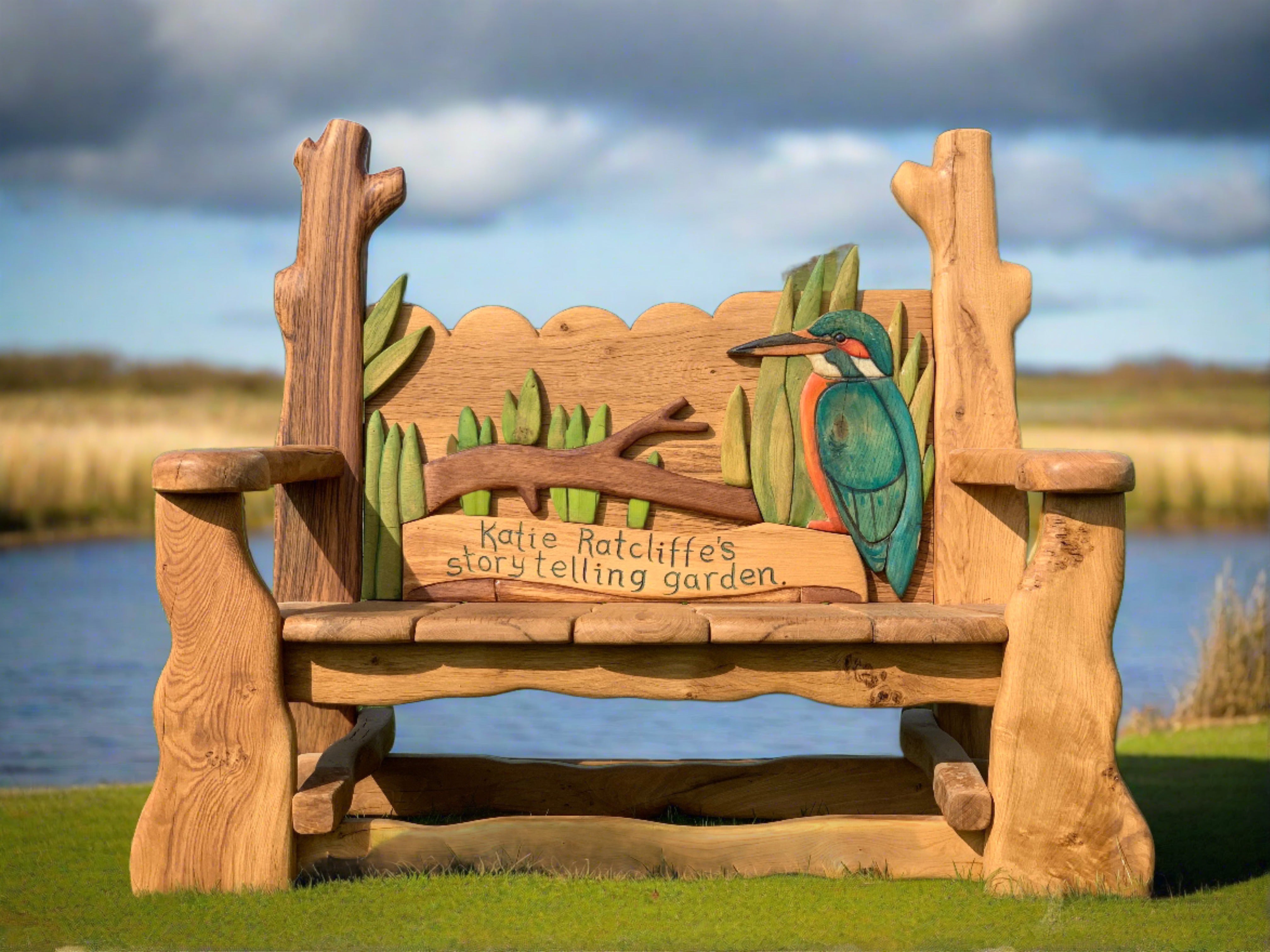 Handcrafted wooden memorial bench by Free Range Designs, adorned with a beautifully carved kingfisher and reeds, dedicated to Katie Ratcliffe’s storytelling garden, providing a tranquil and artistic seating area for remembrance and storytelling.